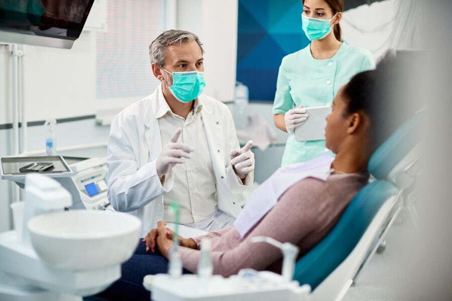 Male dentist wearing face mask while communicating with African American woman during dental appointment at dentist's office.