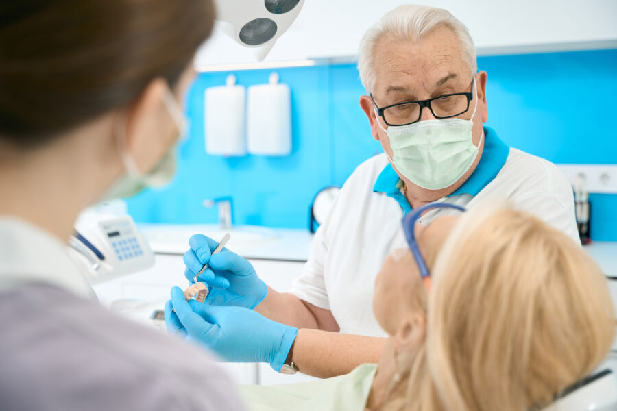Close up portrait of handsome man doctor in glasses is holding teeth model and metal tool while discussing with patient in dental clinic